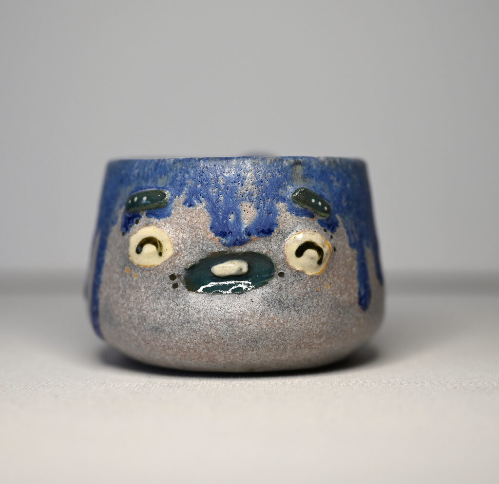 Front view of But WHY? by Fatin Rahmouni, handmade ceramic Monstermug with blue dripping glaze, wide eyes and a puzzled expression.